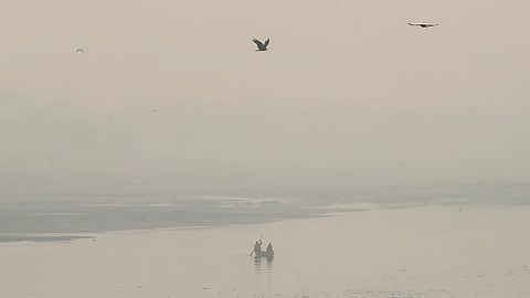 A scene of two people in a small boat on a misty river, with several birds flying above. The image depicts delhi's hazardous air pollution.