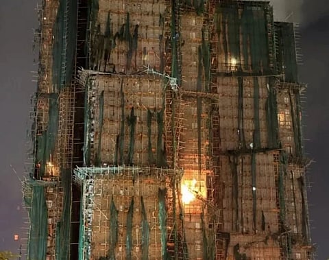 Image of Hong Kong fire. The building is covered in scaffolding and green netting, illuminated by glowing orange lights at night. The atmosphere is dramatic and intense.