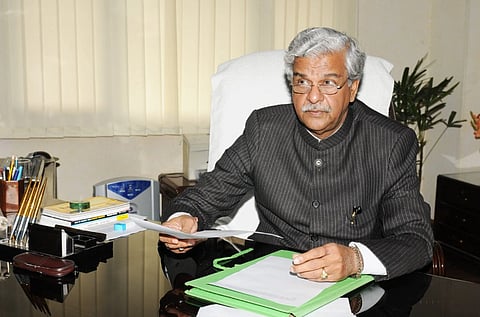 image of Former Union Minister Shriprakash Jaiswal in a suit. He is sitting at a desk holding papers. Office decor includes plants and supplies.