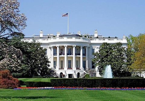 The White House with an American flag atop, surrounded by blooming trees and manicured lawns. A prominent fountain is in the forefront, under a clear blue sky.