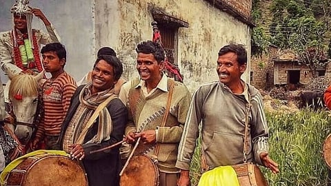 Ceremony in a rural village with three men smiling and playing drums, while people and a horse in the background suggest a festive atmosphere.