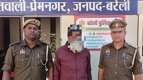 Two police officers stand on either side of a man with a blurred face in front of a building with Hindi text. The atmosphere appears formal.