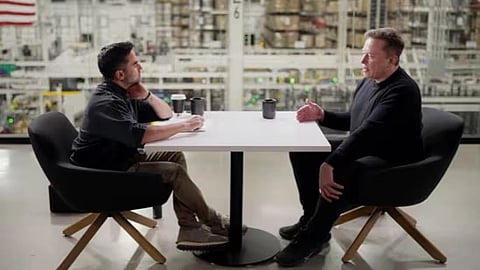 Nikhil Kamath and Elon Musk are engaged in conversation at a table in an industrial setting. The atmosphere is focused and professional, with cups on the table.