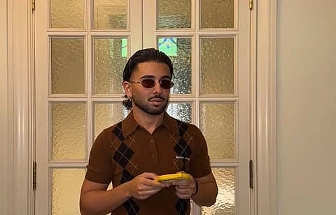 Orry with sunglasses and a brown argyle-patterned shirt holds a yellow phone, standing in front of textured glass doors, conveying a relaxed, stylish vibe.