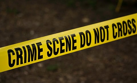 Yellow crime scene tape with bold black text stating "CRIME SCENE DO NOT CROSS" stretches across a blurred outdoor background, conveying a serious tone.