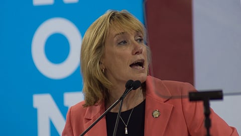 Maggie Hassan in a coral blazer speaks passionately at a podium against a backdrop of blue and white text. The setting suggests a political or formal event.
