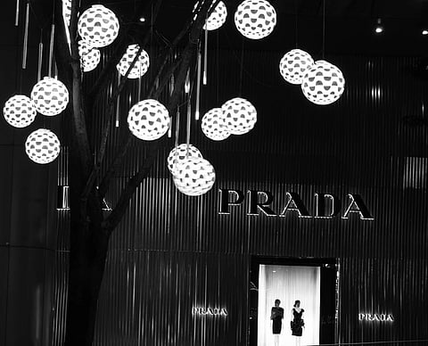 Black and white photo of a Prada storefront, illuminated by spherical, patterned lights hanging from a tree, evoking a chic and elegant mood.