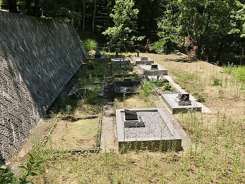 An Islamic graveyard with a wall to the left and trees in the background. There are graves organised in two rows from the wall.