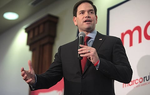 Image of state secretary Marco Rubio in a suit speaking into a microphone at an indoor event. He gestures with one hand, exuding confidence. Partial sign with text is visible behind him.