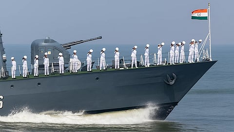 The Indian Navy in their white uniforms stand in formation on the ship's deck, saluting the Indian flag waves at the bow against a blue ocean backdrop.