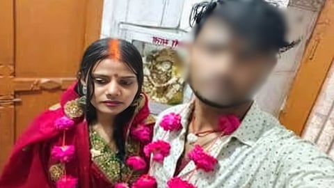 Poonam in traditional Indian attire with a red saree and floral garland stands beside a man with blured face wearing a light shirt. The setting is indoors with a wooden door in the background. The mood appears solemn.