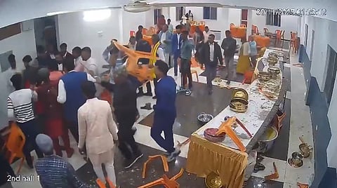 A chaotic scene at an indoor event shows people in formal attire engaged in a fight, throwing orange chairs. Tables are overturned, with food and utensils scattered. Tension and disorder are evident.