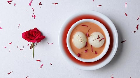 A white bowl with two round, white rasgullas in orange syrup, garnished with saffron and rose petals. A red rose and scattered petals on the left.