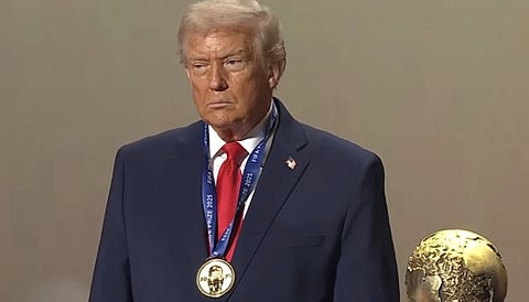 Image of President Donald Trump with Fifa peace prize and medal on his neck. He is looking towards his right and is wearing his sharp blue suit with his red tie. On the bottom right side of his image, a gold trophy is seen with a globe on top of it.