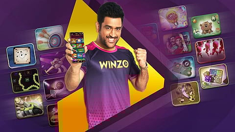 MS Dhoni smiling and pumping his fist while standing in a WinZO jersey. He is holding a phone with the app open. The background is a graphic with WinZO apps.