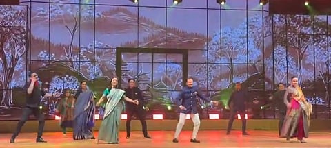 A group of people energetically dancing on stage in traditional Indian attire, with a scenic backdrop showing a mountainous and forested landscape.