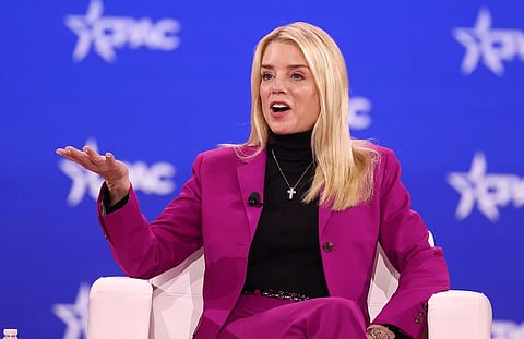 Image of US Attorney General Pam Bondi in a vibrant purple suit speaks animatedly on stage at a conference, gesturing with one hand. The blue backdrop features a star logo and the acronym "CPAC."