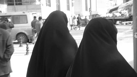 Two people wearing black burqas walk on a sunlit sidewalk in a busy urban setting. Vehicles and other pedestrians are visible in the background.