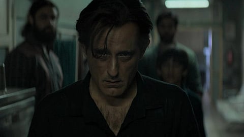 Actor Akshaye Khanna with disheveled hair and a dark shirt stands in a dimly lit hallway, flanked by three blurred figures in the background