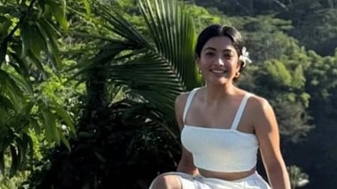 Image of actress rashmika Mandanna in a white dress with greenery behind her. She has a white flower on her hair.