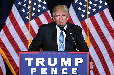 Image of President trump standing at a podium with "TRUMP PENCE" signage. He wears a suit and tie, against a backdrop of multiple American flags,