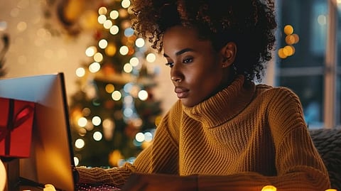 A woman in a cozy room types on a laptop, surrounded by festive Christmas lights and wrapped gifts. She wears a warm sweater, conveying a focused and serene mood.