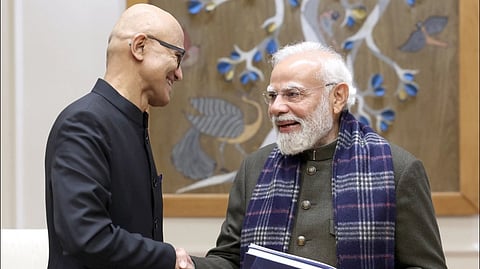 Microsoft CEO Satya Nadella in a dark suit and glasses shakes hands with Prime minister Narendra Modi who is in a green coat and patterned scarf. They are smiling warmly in a formal setting.