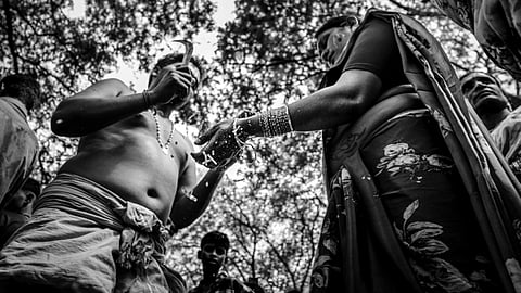 Black and white image of a cultural ritual, featuring a man breaking the bangles of a trans women, surrounded by trees and onlookers.