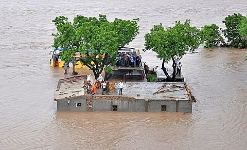 Flood Affected Areas of Amreli District Gujarat India on 24 in June 2015. A building and a bus in the middle of a flood, with people stuck on the roof of the building and trees nearby.