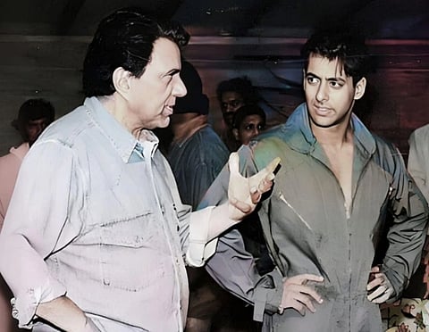 Image of Dharmendra and Salman Khan standing. they are engaging in a conversation. Onlookers are blurred in the background, creating a focused, intense atmosphere.
