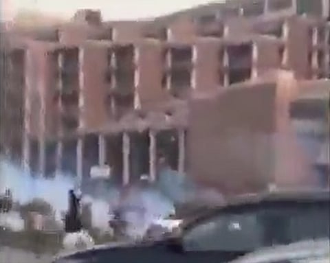 Blurry image depicting a multi-story brick building. Smoke or mist billows in front, obscuring details. A dark vehicle is partially visible in the foreground.