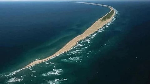 Aerial view of the narrow, sandy Sable Island stretching across the ocean, connecting two landmasses. Waves gently crash on both sides under a clear sky.