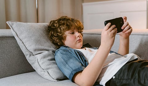A young boy with curly hair lounges on a gray sofa, intently looking at a smartphone. He wears a denim shirt, conveying a relaxed mood indoors.