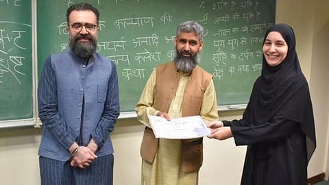 Three people stand in front of a chalkboard with Hindi writing. A smiling woman in a hijab receives a certificate from a man in a vest. The mood is celebratory.