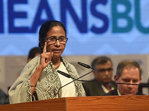 West Bengal CM, Mamata Banerjee in a patterned shawl and glasses speaks at a podium with a serious expression, gesturing with her hand.