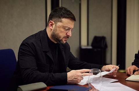 Ukrainian President Volodymyr with glasses, wearing a dark jacket, is seated at a desk, intently reading documents. A notepad and glass are nearby, conveying focus.