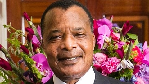 President Denis Sassou-Nguesso in a suit and tie smiles warmly in front of vibrant pink and purple flowers, conveying a sense of joy and elegance.