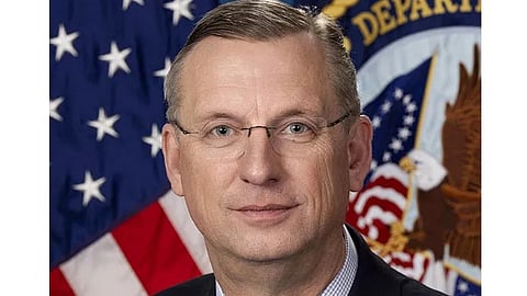 Portrait of US Veterans Affairs Secretary Doug Collins in a suit with glasses, set against a backdrop featuring the American flag and a governmental emblem. The mood is formal and authoritative.