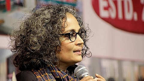 Arundhati Roy with glasses speaks into a microphone, wearing a scarf and black top. A blurred sign with "ESTD." is in the background.