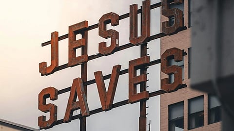 Rustic metal sign reading "JESUS SAVES" mounted high on a building against a cloudy sky, conveying a vintage and spiritual atmosphere.