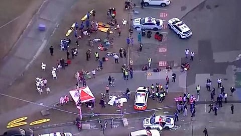 Aerial view of an emergency response scene with multiple police cars, officers, and people gathered. Tents and equipment are visible, conveying urgency.