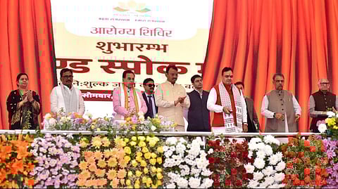Rajasthan CM Bhajan Lal Sharma along with other Ministers standing on stage as he launches the new scheme with vibrant red curtains. They're positioned behind a facade adorned with an array of colorful flowers.