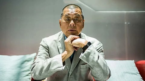 Hong Kong Media Tycoon Jimmy Lai in a gray blazer sits on a red sofa, resting his chin on his clasped hands. He appears thoughtful, with a neutral expression. Gray cushions are beside him.