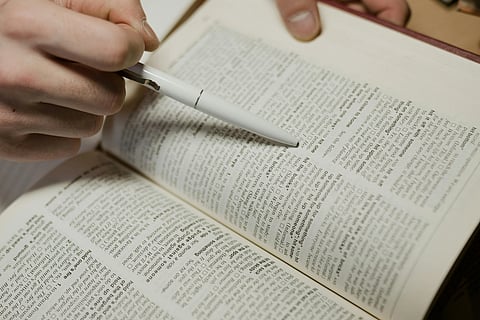 A person holds a white pen above an open dictionary, pointing at a word. The scene conveys focus and learning, with text clearly visible on the pages.