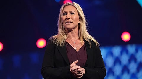 Marjorie Taylor Greene with long blonde hair speaks on stage, wearing a dark blazer. The backdrop features abstract patterns and red stage lights, creating a focused atmosphere.
