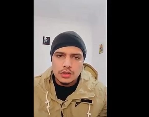 Australian-Based Haryanvi Rapper Dhanda Nyoliwala wearing a black beanie and a beige jacket looks directly at the camera. The background is a plain white wall with a small picture on it.