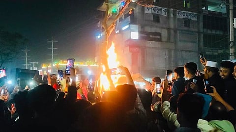 Violent protest led by a large crowd surrounding a burning fire. Many people hold up phones to capture the fiery scene.
