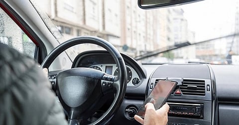 A person drives in rainy weather, holding a smartphone with one hand and the steering wheel with the other. The windshield wipers are active, creating a risky situation.