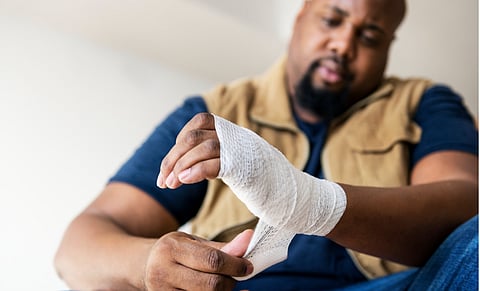 A man attentively wraps a bandage around his injured wrist. He appears focused and calm, wearing a blue shirt and brown vest, seated in a relaxed position.