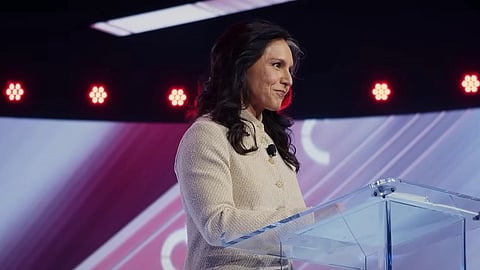 Tulsi Gabbard in a light-colored blazer speaks confidently at a clear podium on stage, with a colorful backdrop and red lights, conveying a professional tone.